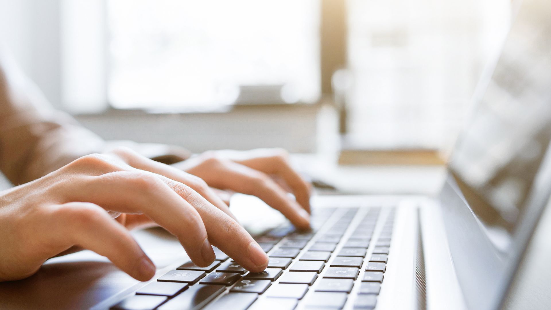 Close-up of hands typing on a laptop keyboard, with soft natural light coming through a window in the background.